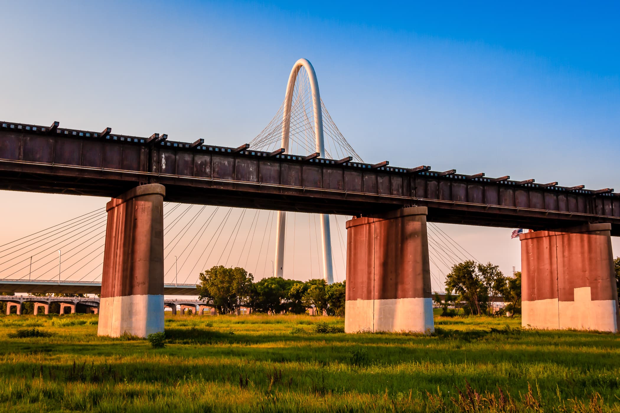 A railroad bridge, in the foreground, is overwhelmed by the massive arch and cables of Dallas' Margaret Hunt Hill Bridge as the sun sets over the DFW Metroplex.