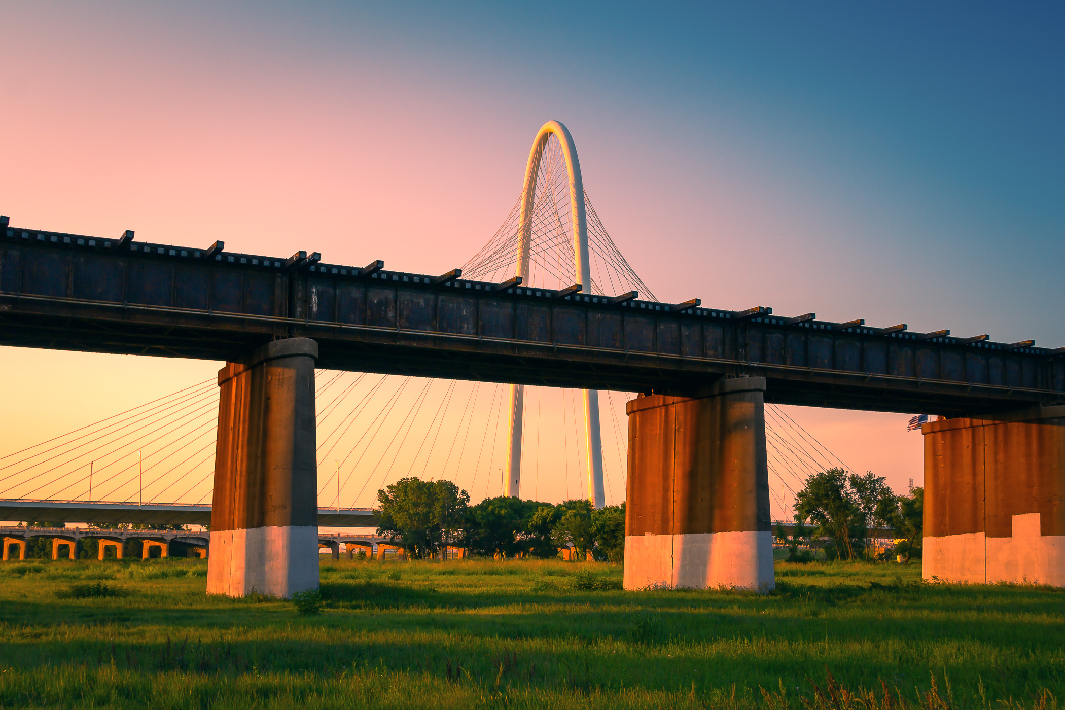 A railroad bridge, in the foreground, is overwhelmed by the massive arch and cables of Dallas' Margaret Hunt Hill Bridge as the sun sets over the DFW Metroplex.