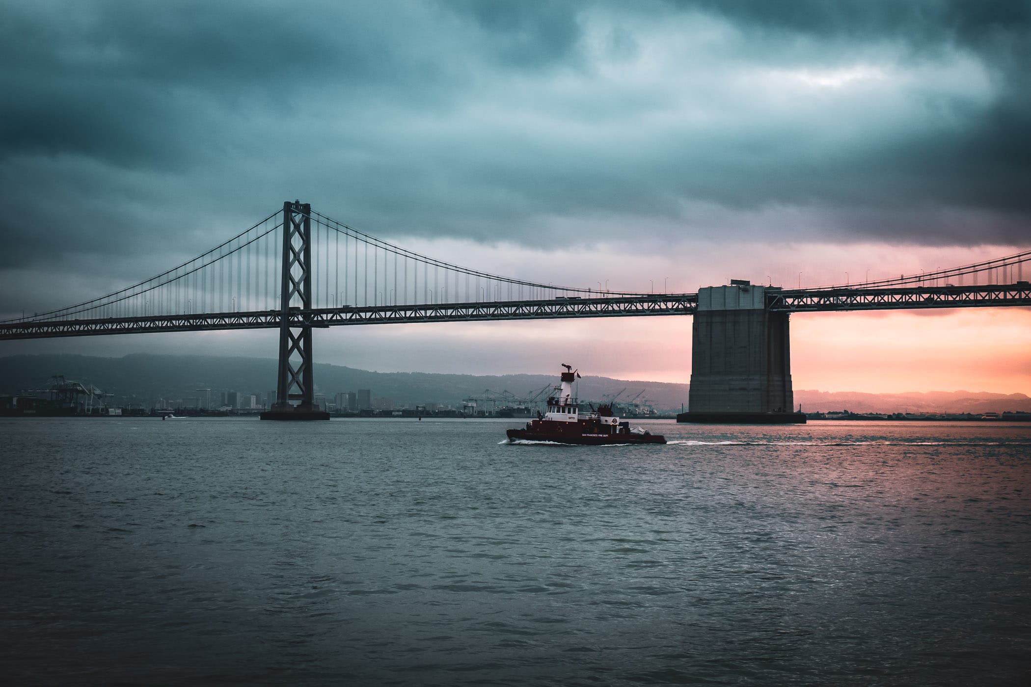 A San Francisco Fire Department boat traverses San Francisco Bay as the Bay Bridge rises into the gloomy sky of morning.