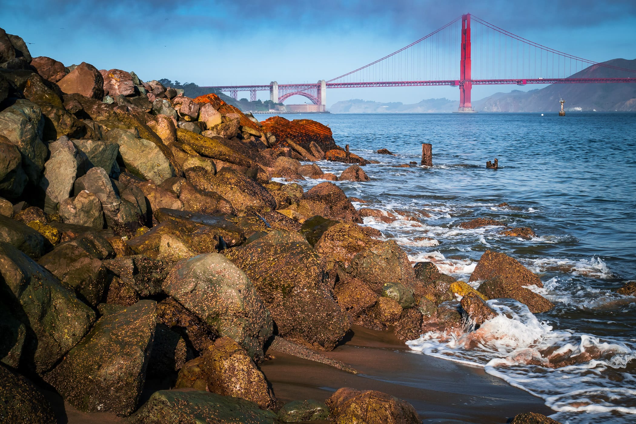 The San Francisco Bay surf laps at rocks on the beach at Crissy Field as the Golden Gate Bridge rises in the distance.