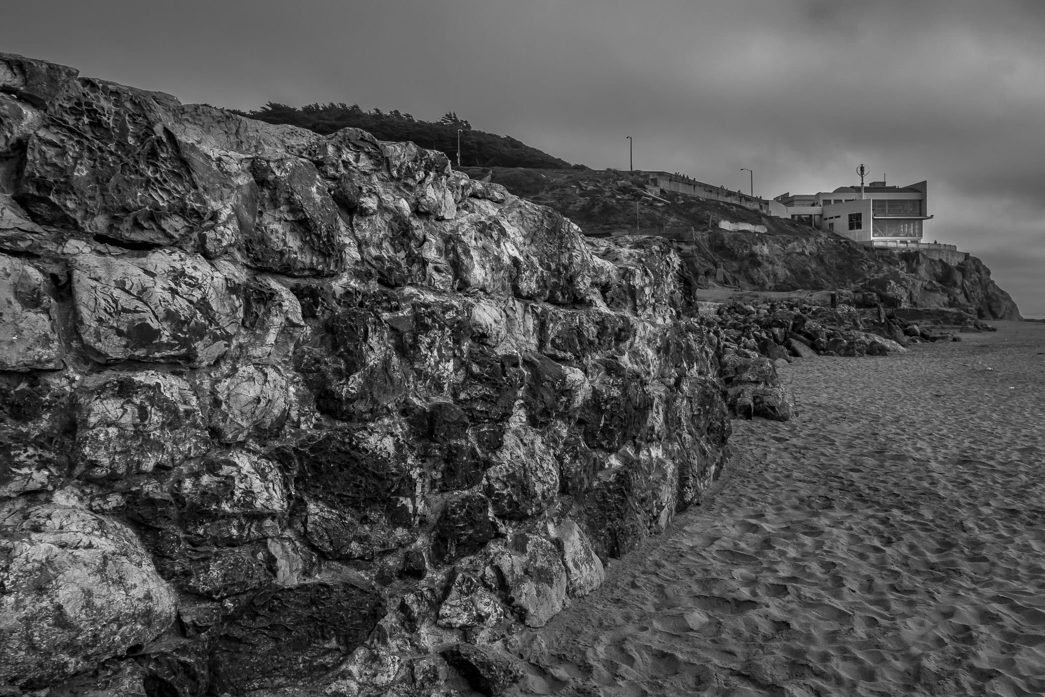 A rock wall—part of the ruins of the Sutro Baths—leads towards the modern wing of the historic Cliff House restaurant on the beach at Lands End, San Francisco.